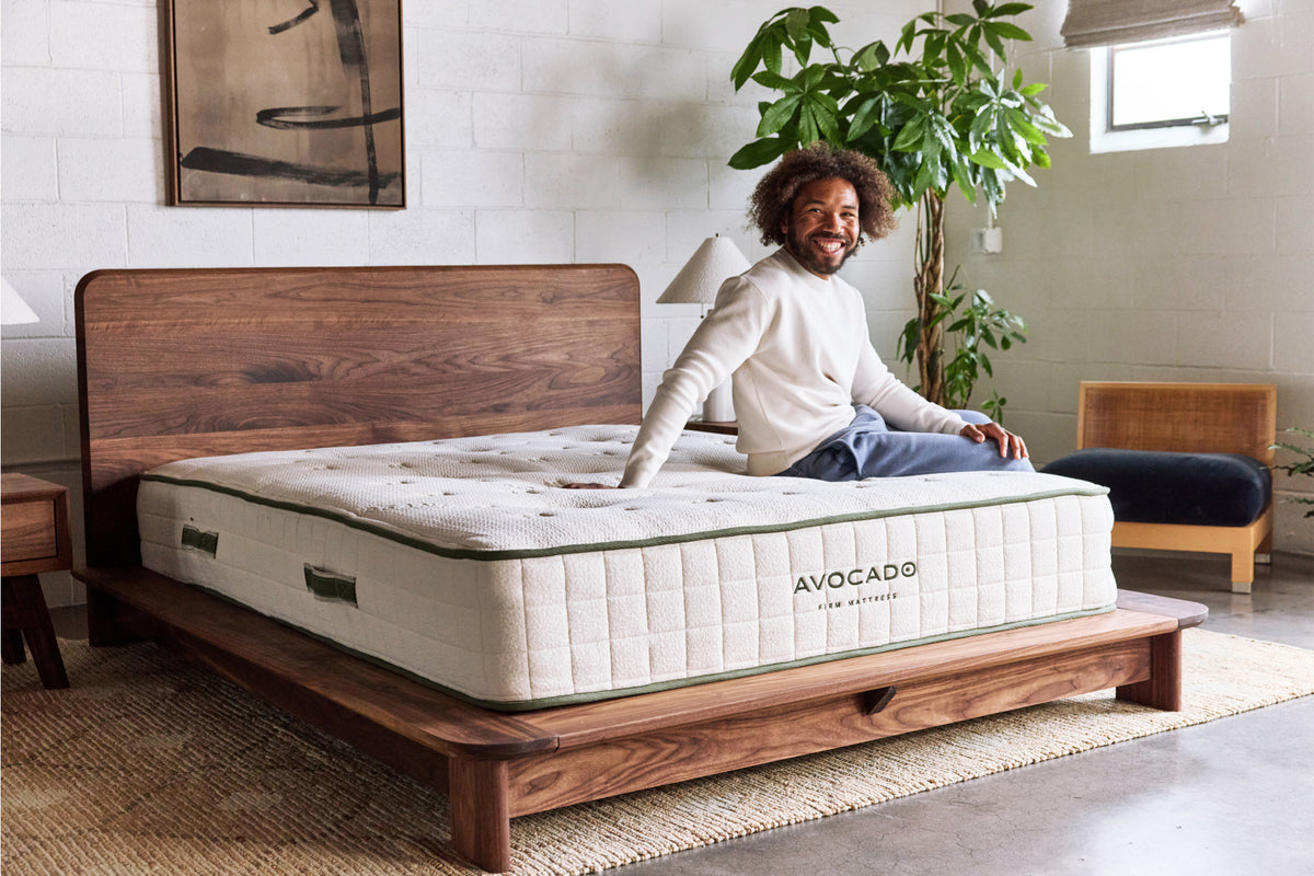 A man sits on an Avocado mattress on a wooden bed frame in a modern, sunlit bedroom. The mattress has a quilted cream-colored top and side with green trim, and the brand name 'AVOCADO' is clearly visible on the side panel.