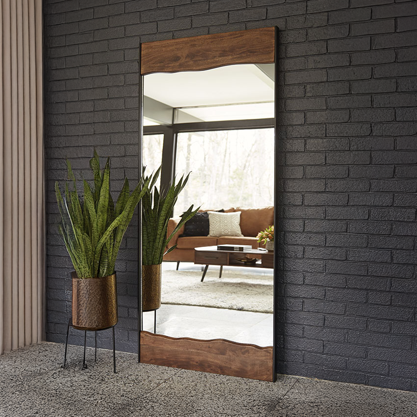 Full-length mirror with wooden frame leaning against a dark brick wall, reflecting a living room.