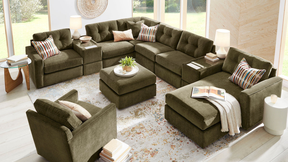 An olive green sectional sofa set arranged in a bright, modern living room with large windows, light wood floors, and a patterned area rug. The set includes an L-shaped sectional, a matching armchair, a central ottoman, and striped accent pillows.