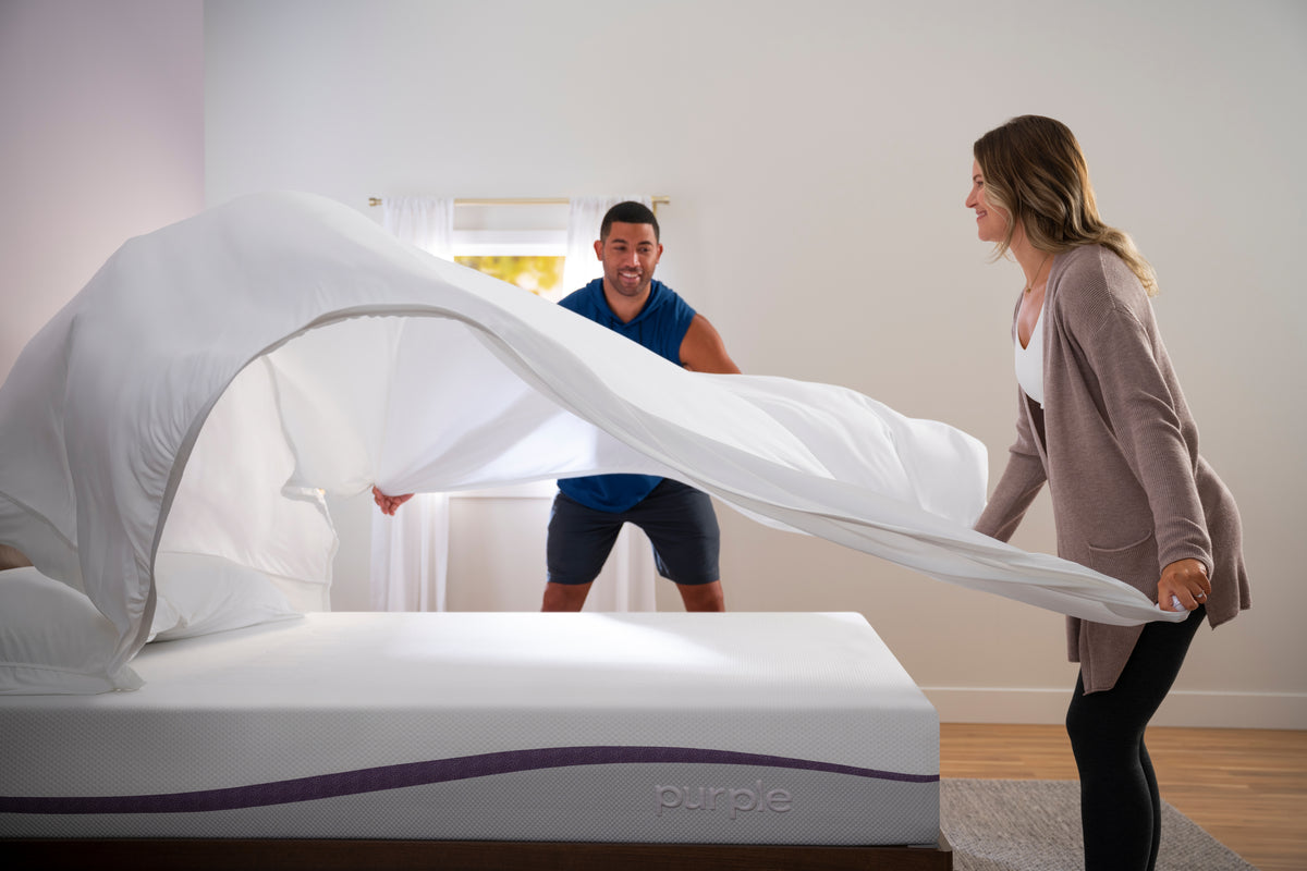 Two people unrolling a white sheet on a bed with a 'Purples' brand logo.